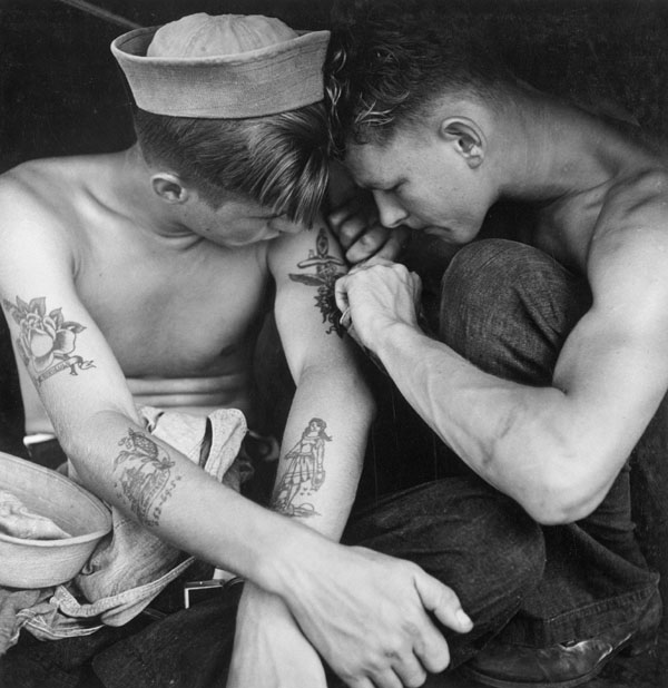 A black and white photo of two young sailors tattooing one another

